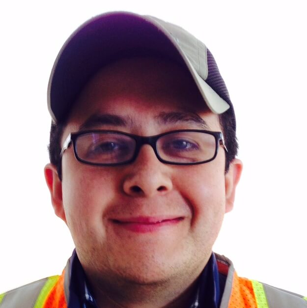 A person wearing glasses, a gray cap, and an orange safety vest is smiling at the camera against a white background, ready for their next Antarctic Logistics and Expeditions adventure in Antarctica.