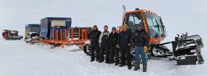 Five people in winter gear stand in front of an orange snow vehicle with treads, attached to sledges carrying equipment, on a snowy Antarctic landscape under an overcast sky, likely part of an Antarctic Logistics and Expeditions journey.