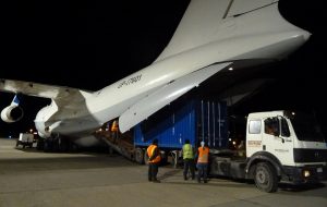 Three workers in reflective vests stand by a large cargo truck as a shipping container is unloaded from the open cargo bay of an airplane at night on an airport tarmac, supporting Antarctic Logistics and Expeditions for operations near the South Pole.