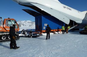 People unload a large blue shipping container from an open cargo plane onto snowy ground at the South Pole, with mountains in the background and a tracked vehicle nearby. Several workers in winter gear handle this Antarctic Logistics and Expeditions task.
