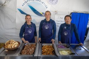 Three people in matching blue aprons stand behind a buffet table with trays of food and sliced bread inside a tent marked “Antarctic Logistics and Expeditions,” set up at the South Pole, with a clock on the wall.