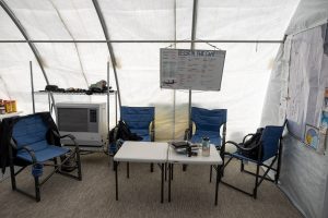 Four blue camping chairs arranged around two white folding tables inside a tent in Antarctica. The tables hold books, a mug, and other items. A whiteboard with notes hangs on the wall, and a large South Pole map is posted on the right.