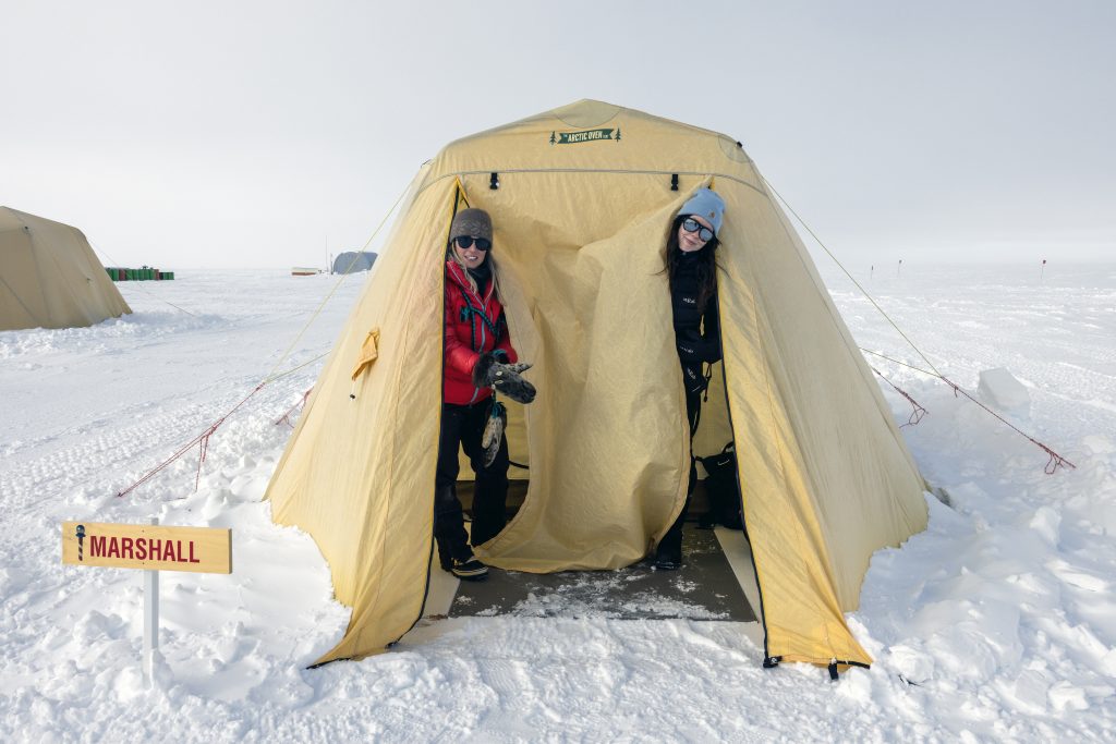 Two people in winter clothing stand smiling at the entrance of a yellow tent in snowy Antarctica. A sign reading Marshall is beside the tent, with other tents from Antarctic Logistics and Expeditions visible in the background.