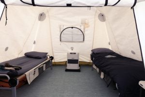 Interior of a tent in Antarctica with two single beds on opposite sides, cozy bedding, and a centrally placed heater by the back wall; sunlight streams through a window behind the heater. Experience comfort with Antarctic Logistics and Expeditions.