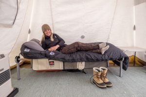 A woman in winter clothing relaxes on a cot inside a tent in Antarctica. She is smiling, lying on a sleeping bag with boots placed neatly beside her and a table in the background, ready for another day with Antarctic Logistics and Expeditions.