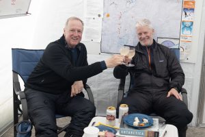 Two men in winter gear sit in a tent near the South Pole, smiling and toasting drinks. Behind them are maps and notes on the wall. On the table are beers, cheese, bread, books, and a plate titled Antarctica.