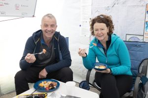 A man and a woman sit indoors on camping chairs, smiling at the camera while eating from blue plates. Both wear outdoor jackets; a map and notes—possibly detailing an Antarctica trip with Antarctic Logistics and Expeditions—are on the wall behind them.