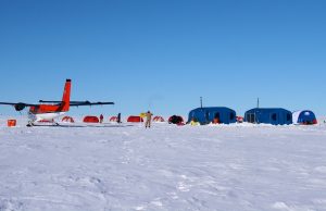 A small red plane is parked on a snowy field in Antarctica near an expedition camp, where red and blue tents and shelters from Antarctic Logistics and Expeditions stand under a clear blue sky.
