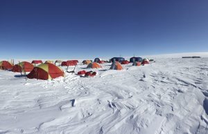 Rows of colorful tents, mostly red and yellow, are set up on a snowy, windswept landscape under a clear, deep blue sky—an Antarctica expedition camp supported by Antarctic Logistics and Expeditions in a remote polar region.