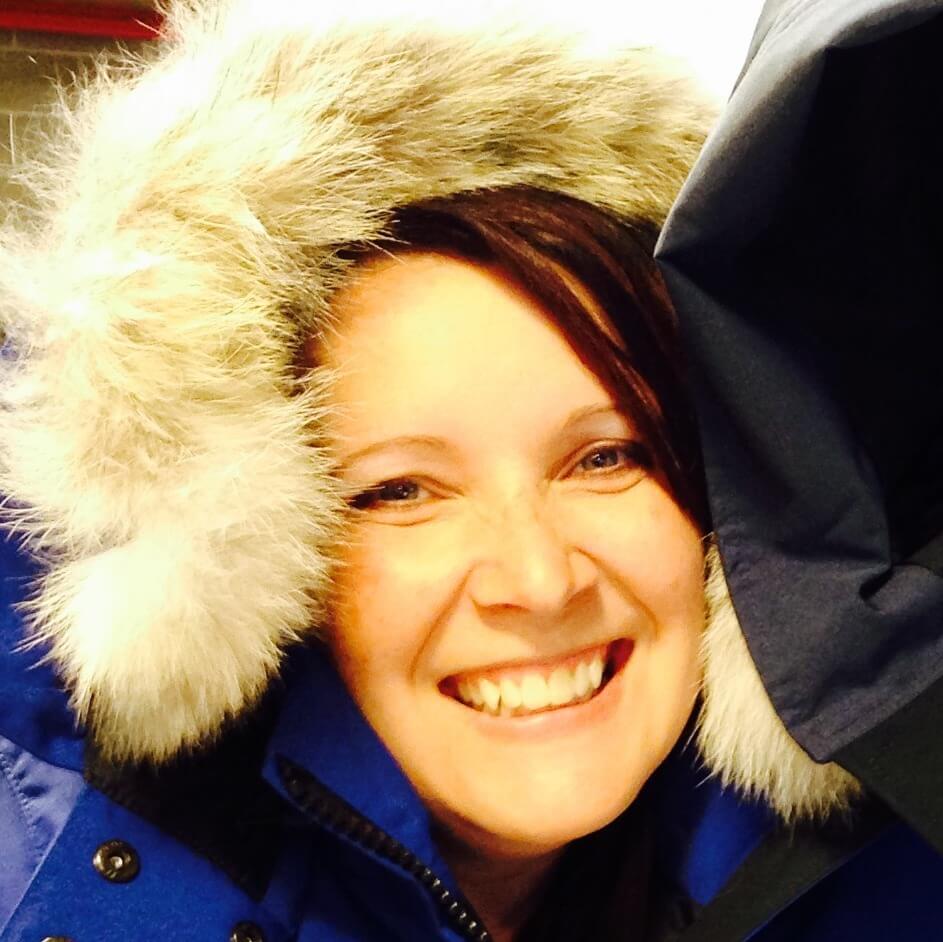 A smiling person wearing a blue winter coat with a furry hood, standing indoors. The brightly lit, close-up photo captures their joyful expression—ready for an Antarctic Logistics and Expeditions journey to Antarctica.