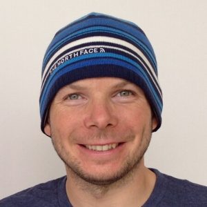 A smiling man wearing a blue, white, and black striped The North Face beanie and a blue T-shirt stands against a plain white background, ready for his next Antarctica adventure with Antarctic Logistics and Expeditions.