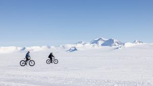 Two people ride fat-tire bikes across a vast, snowy landscape in Antarctica, with distant snow-covered mountains under a clear blue sky.