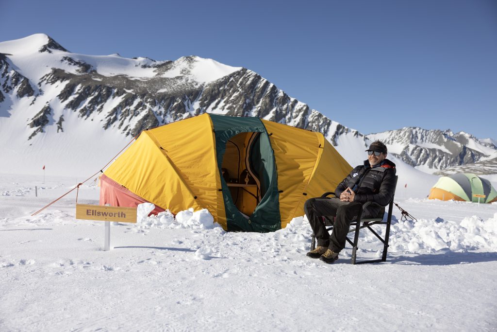 A person in winter clothing sits on a folding chair outside a yellow and orange tent on a snowy Antarctic landscape, with snow-covered mountains in the background and an "Ellsworth" sign, part of an Antarctic Logistics and Expeditions camp.