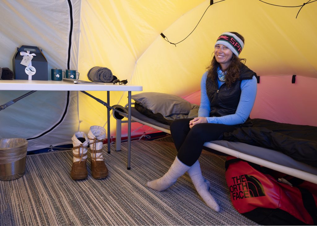 A woman in winter clothes and a beanie sits on a cot inside a brightly lit tent in Antarctica. Nearby, winter boots, a red North Face bag, and camping gear add to the organized, cozy setting—perfect for an Antarctic Logistics and Expeditions adventure.