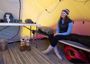 A woman wearing winter clothes sits on a cot inside a yellow camping tent in Antarctica. Next to her is a table with mugs, a teapot, and rolled towels. Winter boots and a red North Face bag are on the floor beside her.