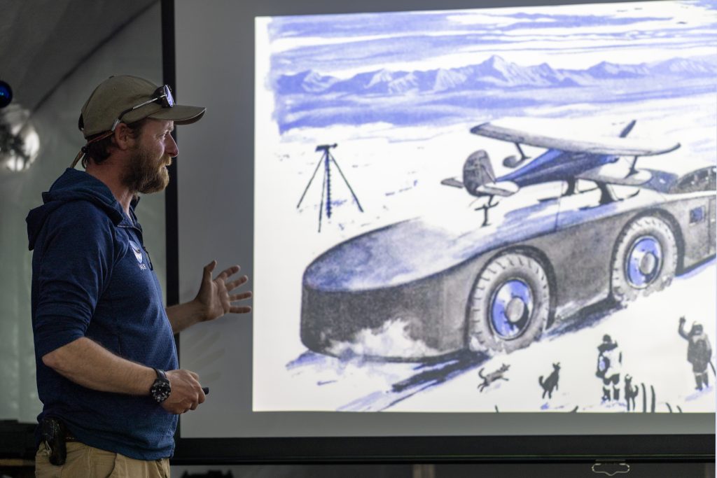 A man in a cap and sunglasses gestures while presenting a projected black-and-white sketch of a futuristic vehicle with an airplane on top, set in a snowy South Pole landscape with mountains in the background.