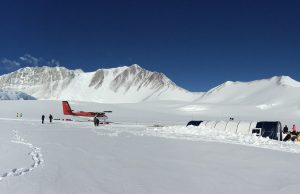 A red airplane operated by Antarctic Logistics and Expeditions is parked on a snowy field near a large white tent with several people walking around. Snow-covered mountains and a clear blue sky form the stunning backdrop in Antarctica.