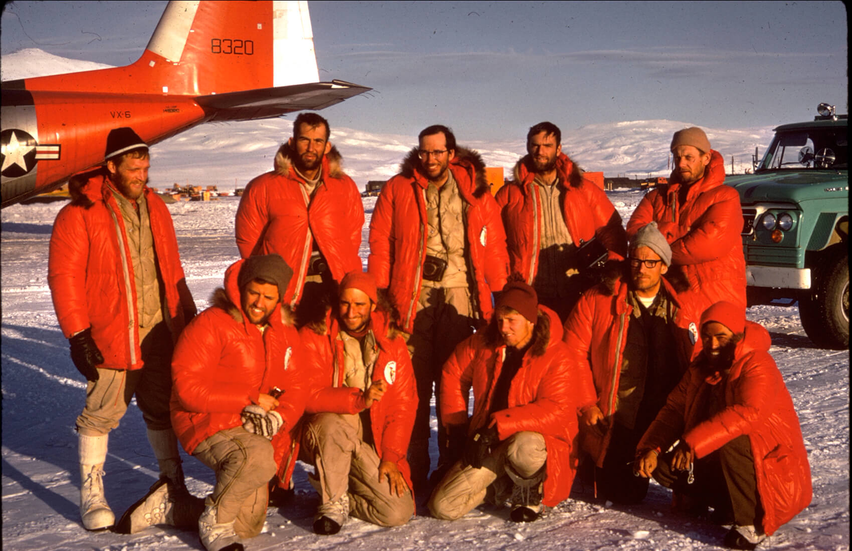 A group of men in red parkas pose for a photo on snowy ground near an aircraft and green vehicle, backed by mountains. They appear to be part of an Antarctic Logistics and Expeditions team in remote Antarctica.