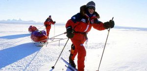 Two people in red snowsuits ski across a snowy, icy Antarctic landscape, each pulling sleds loaded with gear. The sky is clear and bright, and the figures are smiling, capturing the spirit of an adventurous expedition supported by Antarctic Logistics and Expeditions.