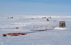 Cargo pallets and orange parachutes scattered across a vast, snowy landscape under a clear blue sky, likely indicating an Antarctic Logistics and Expeditions airdrop operation in Antarctica or near the South Pole.