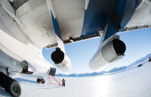 A person stands near the rear landing gear of a large airplane on an icy surface in Antarctica, with snow-covered mountains and a clear blue sky behind them. The image is taken from beneath the aircraft's wings, highlighting Antarctic Logistics and Expeditions.