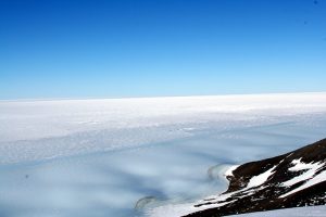 A vast icy landscape under a clear blue sky, with a snow-covered slope and patches of exposed rock in the foreground, leads to a flat expanse stretching to the horizon—capturing the remote beauty of Antarctica near the South Pole.