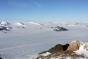 Vast icy landscape with snow-covered mountains in the distance, flat frozen terrain in the foreground, and some exposed rocky peaks under a clear blue sky—an iconic view of Antarctica near the South Pole.