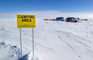 A yellow sign reading Camping Area, Worlds Southernmost Resort stands on snowy ground in Antarctica, with tents and shelters in the background under a clear blue sky—part of Antarctic Logistics and Expeditions’ remarkable offerings.