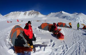 People in red and yellow jackets stand among orange tents on snow in Antarctica, surrounded by snowy mountains. Two small planes, part of Antarctic Logistics and Expeditions, are visible under a clear blue sky.