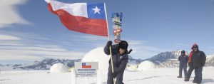A person in winter gear plants a large Chilean flag in snowy Antarctic terrain near a signpost, with dome tents and two others standing in the background; snow-covered mountains are visible under clear skies.