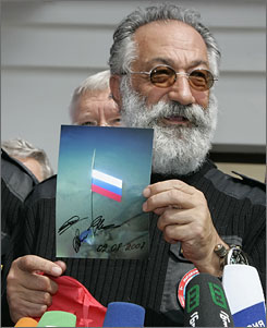 A man with gray hair and a beard wears sunglasses and holds up a signed photo of a Russian flag planted underwater in Antarctica. He stands behind several colorful microphones at what appears to be an Antarctic Logistics and Expeditions press event.