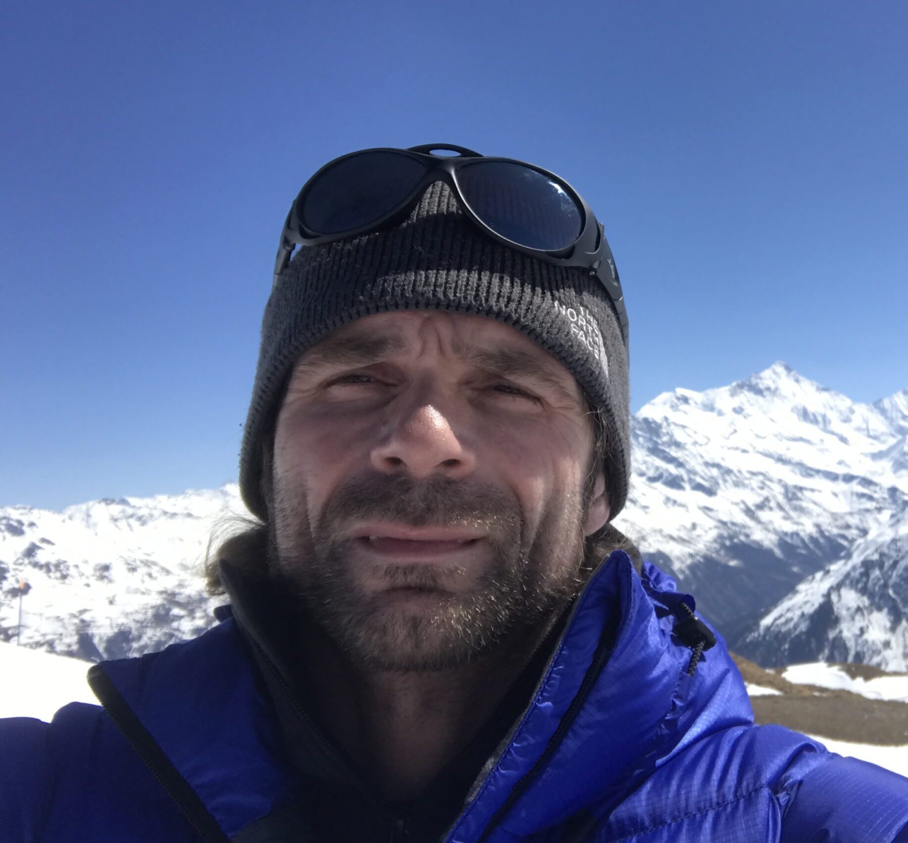 A man wearing a gray beanie, sunglasses on his head, and a blue jacket takes a selfie outdoors with snow-covered mountains and a clear blue sky in the background near the South Pole during an Antarctic Logistics and Expeditions journey.