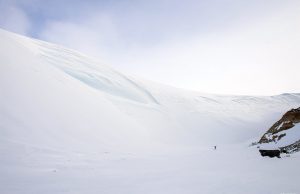 A lone person stands on a vast, snow-covered Antarctic landscape beside a steep, icy slope under a pale sky, with a small, dark rocky outcrop on the right—a scene reminiscent of Antarctic Logistics and Expeditions journeys.