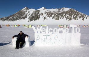 A person in winter clothing sits on a snow chair next to a large Welcome to Antarctica sign made of snow, with Antarctic Logistics and Expeditions tents and snow-covered mountains in the background.