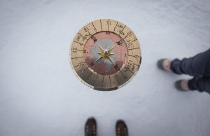 A top-down view of a brass sundial on snowy ground in Antarctica, with two pairs of feet—one in brown shoes at the bottom and another in gray shoes at the right edge—hinting at Antarctic Logistics and Expeditions support.