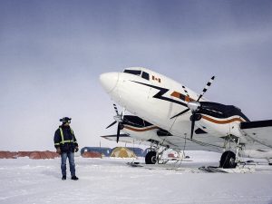 A person in winter gear stands beside a white propeller airplane with a Canadian flag, parked on snowy ground near several colorful tents under a cloudy sky, ready for Antarctic Logistics and Expeditions in Antarctica.