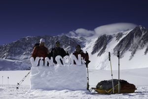 Three climbers in winter gear stand behind a snow sign that reads VINSON, with trekking poles and gear bags nearby. Snow-covered mountains and a blue sky capture the spirit of Antarctica’s remote beauty.