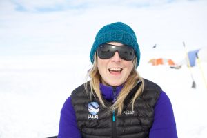 A woman wearing a teal knit beanie, dark sunglasses, and a black puffer vest over a purple top smiles outdoors on a snowy Antarctic landscape with tents and equipment from Antarctic Logistics and Expeditions in the background.
