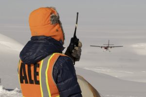 A person wearing a bright orange hood, safety vest, and gloves holds a walkie-talkie while watching a small red plane land on the snowy Antarctic landscape, supporting operations for Antarctic Logistics and Expeditions.