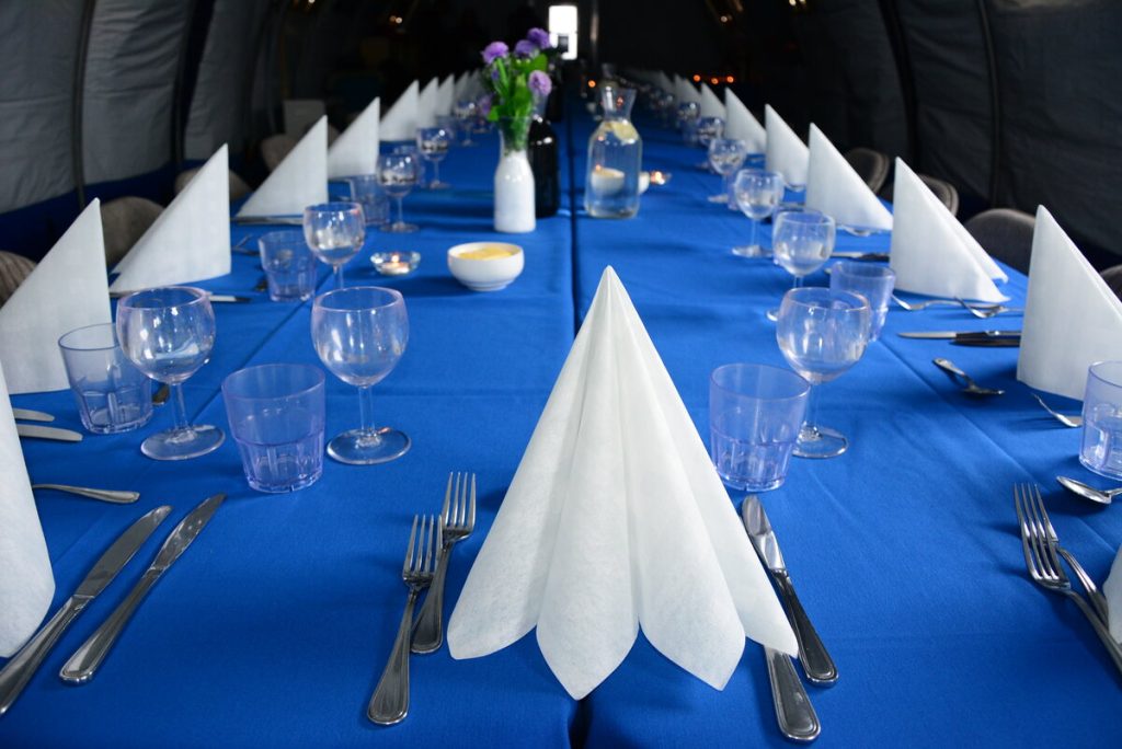 A long table covered with a blue tablecloth is set for a formal meal at Antarctic Logistics and Expeditions, with folded white napkins, clear glassware, silver cutlery, and a vase of purple flowers as a centerpiece.