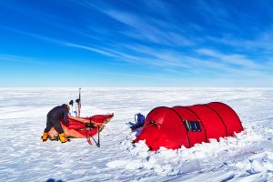 Two people set up camp on a snowy, flat Antarctic landscape next to a bright red tent under a blue sky. Snow gear and camping equipment are scattered around them, capturing the spirit of exploration at the South Pole.