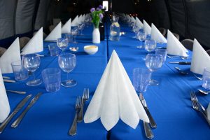 A long dining table covered with a blue cloth, set for a formal meal in a well-lit room, evokes the elegance of an Antarctic Logistics and Expeditions gathering, with folded white napkins, clear glassware, silver cutlery, and small flower vases.