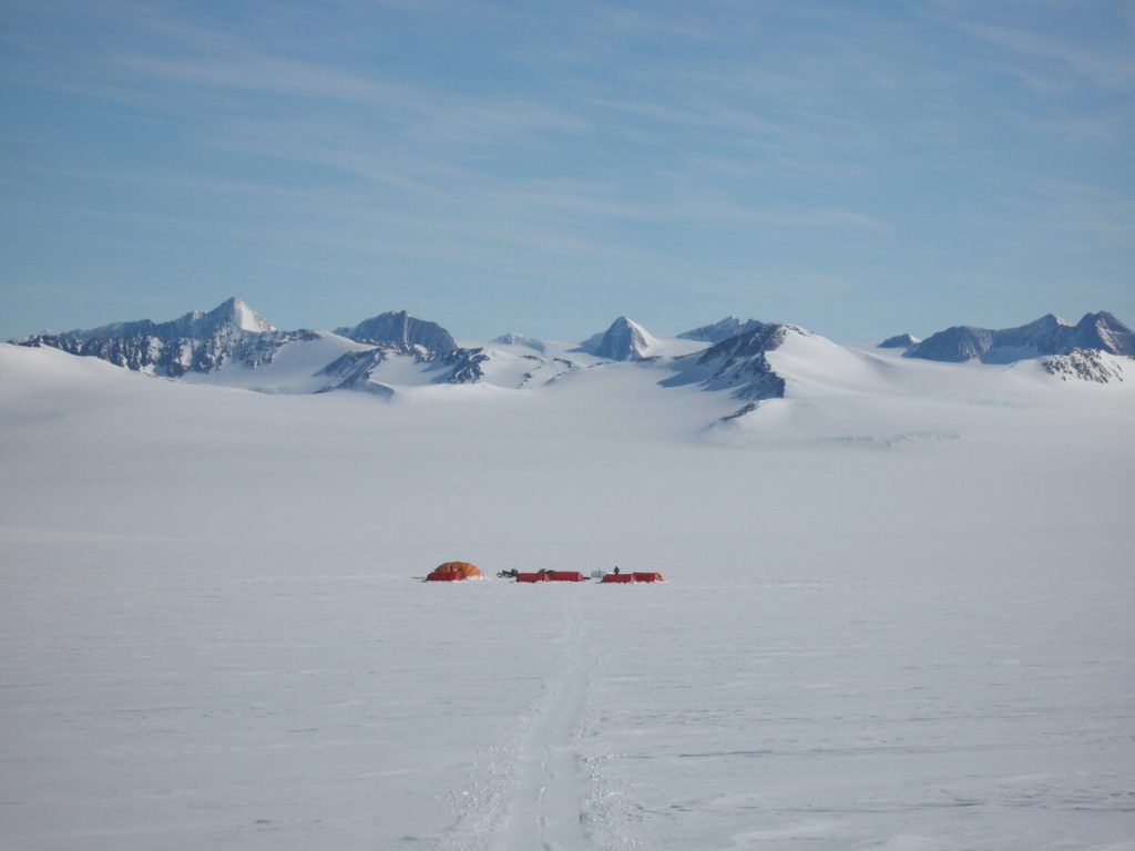 A small cluster of red and orange tents sits on a vast snowy plain in Antarctica, surrounded by snow-covered mountains under a blue sky. Tracks in the snow lead toward the tents, marking an expedition supported by Antarctic Logistics and Expeditions.
