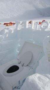 An outdoor toilet made of snow and ice sits in a snowy camp in Antarctica. "Hello" is carved above the structure, with colorful tents from Antarctic Logistics and Expeditions visible on the snow-covered landscape in the background.
