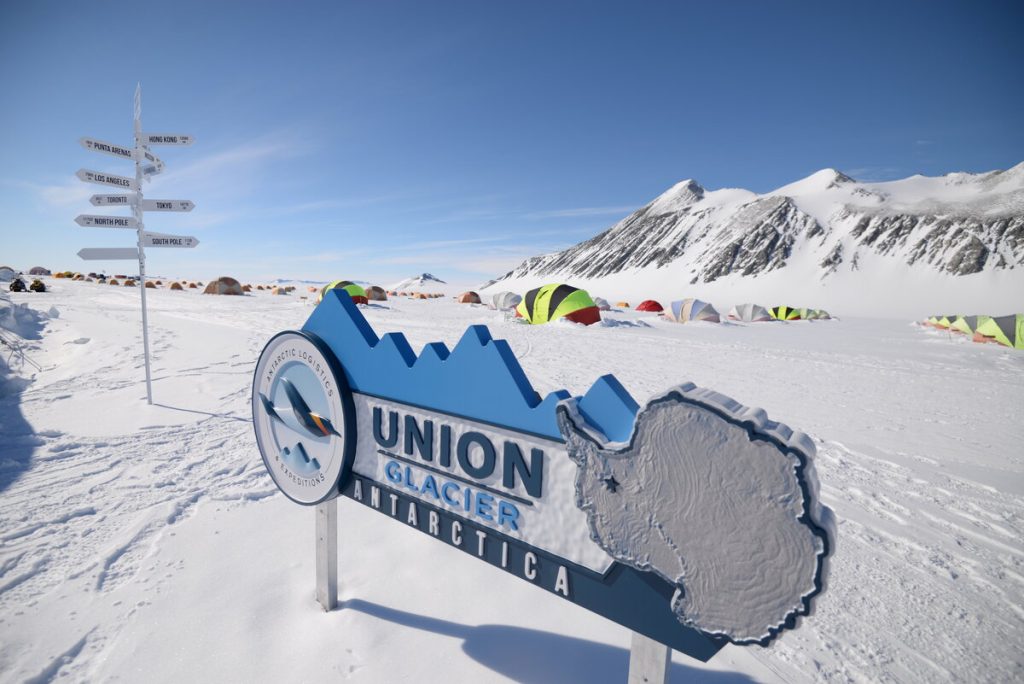 A sign reading Union Glacier Antarctica stands in front of a snowy landscape with colorful tents, distant mountains, and a signpost with multiple directional arrows—an iconic scene supported by Antarctic Logistics and Expeditions under a clear blue sky.