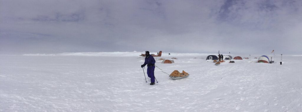 A person in blue cold-weather gear pulls a sled across a snowy Antarctic landscape toward tents and a small red plane in the background, under a cloudy sky.