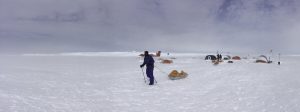 A person in blue cold-weather gear pulls a sled across a snowy Antarctic landscape toward tents and a small red plane in the background, under a cloudy sky.