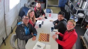 Five people sit around a table inside a tent at the South Pole, smiling at the camera. Playing cards, a chessboard, mugs, and snacks are on the table. The atmosphere appears relaxed and friendly.