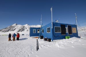 A blue research station stands on a snowy field in Antarctica with solar panels and equipment outside. Four people in winter clothing are talking nearby, with snow-covered mountains and a clear sky in the background.