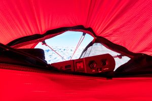 View from inside a bright red tent looking out at snowy mountains near the South Pole, with a portable power station in the foreground—a scene reminiscent of an Antarctic Logistics and Expeditions adventure.